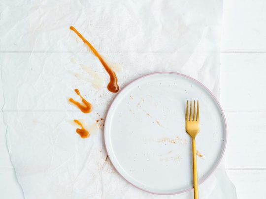 A gold fork resting on an empty white plate on a sunlit table — a quiet image for thinking about how we relate to food and eating
