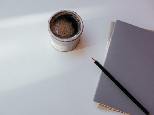 A ceramic cup of coffee, a grey notebook and a pencil on a sunlit desk — a quiet, considered moment for thinking about therapy as a private investment